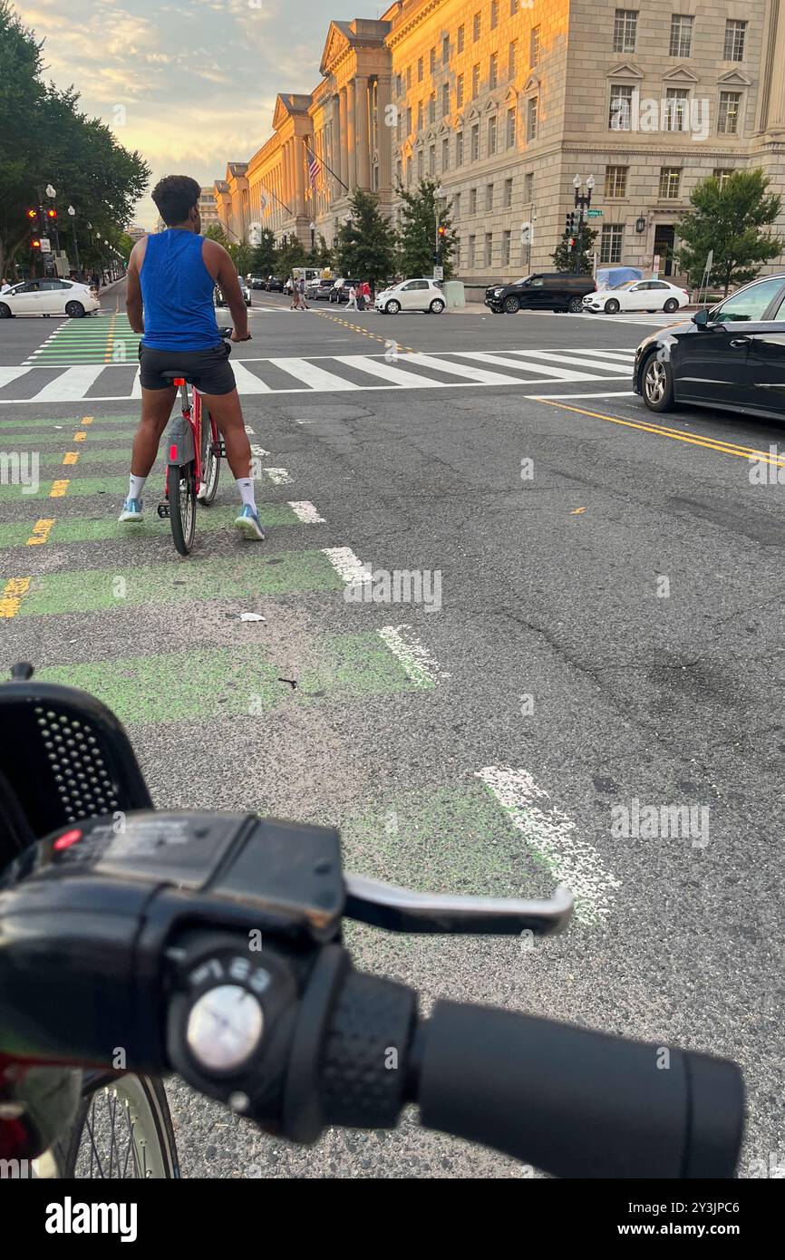 Un uomo in bicicletta sta percorrendo una strada di fronte a un edificio. L'uomo indossa una canotta blu e pantaloncini corti. La strada è fiancheggiata da alberi e t Foto Stock Un uomo in bicicletta sta percorrendo una strada di fronte a un edificio. L'uomo indossa una canotta blu e pantaloncini corti. La strada è fiancheggiata da alberi e t Foto Stock