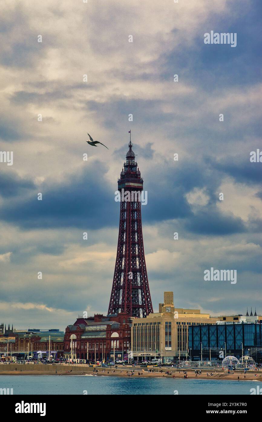 Una vista panoramica di un'alta torre, forse un punto di riferimento, contro un cielo nuvoloso. Il primo piano presenta una spiaggia con persone ed edifici, mentre un uccello vola Foto Stock