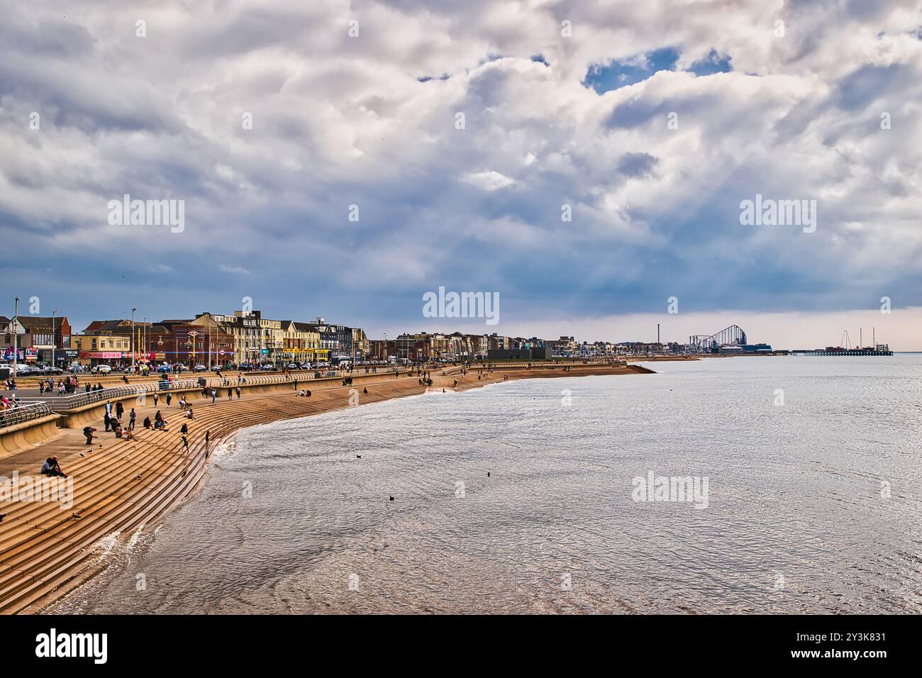 Una vista panoramica di una spiaggia con un litorale sabbioso, persone che camminano e si rilassano. Il cielo è parzialmente nuvoloso con raggi di luce solare che si infrangono. Nella Foto Stock