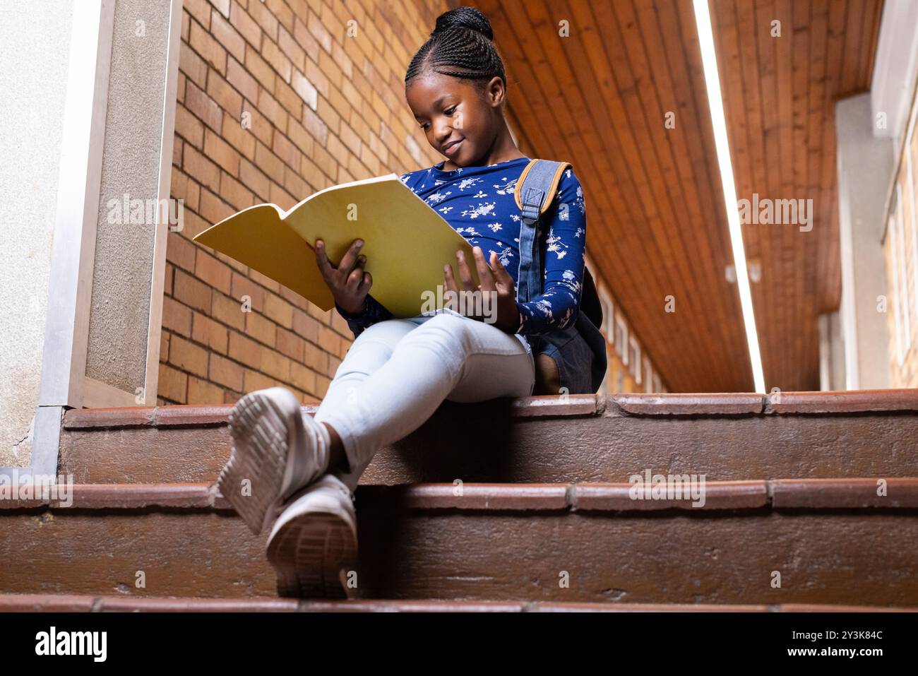 Seduta sulle scale, ragazza afroamericana che legge un notebook con zaino nel corridoio della scuola Foto Stock