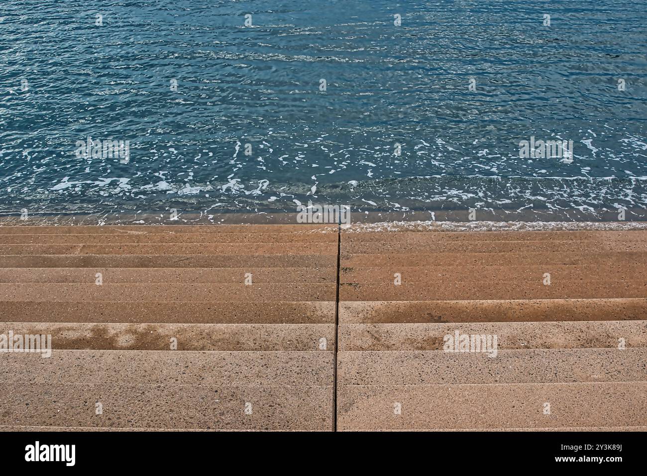 Vista di gradini di cemento che conducono a un calmo corpo d'acqua. I gradini sono asciutti e puliti, con onde delicate che si infrangono sulla base. L'acqua è limpida Foto Stock