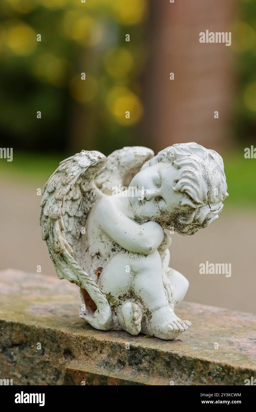 Una piccola statua d'angelo come decorazione funeraria in un cimitero. L'immagine ha uno spazio vuoto per il testo. La statua ha uno sfondo verde estivo. Foto Stock