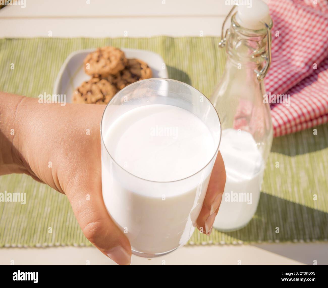 Fotografia di cibo con un bicchiere di latte tenuto in mano a una donna, il biberon di latte e i biscotti stesi sul tavolo sullo sfondo Foto Stock