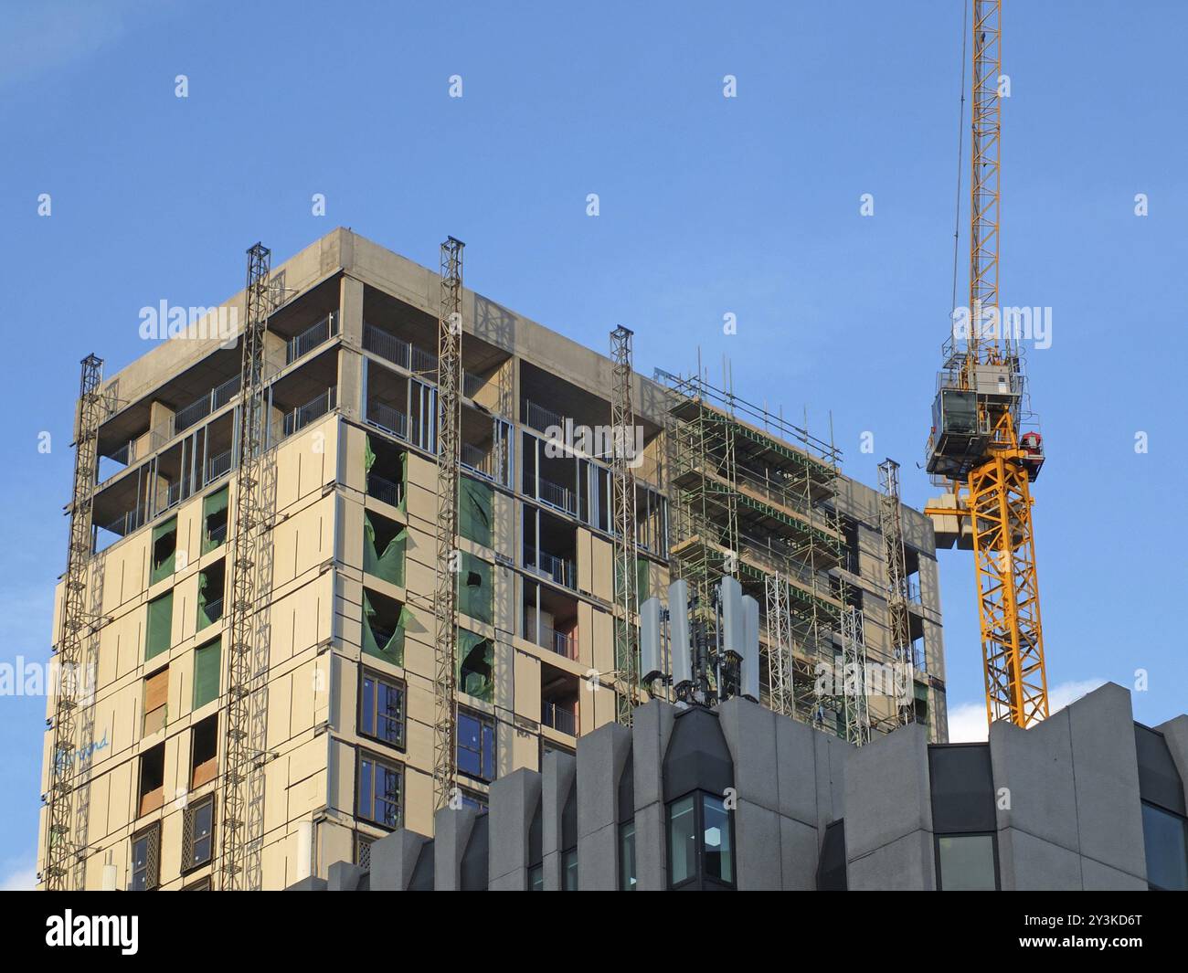 Un'alta gru da costruzione gialla che lavora su un grande edificio in cemento alto con impalcature contro un cielo blu Foto Stock