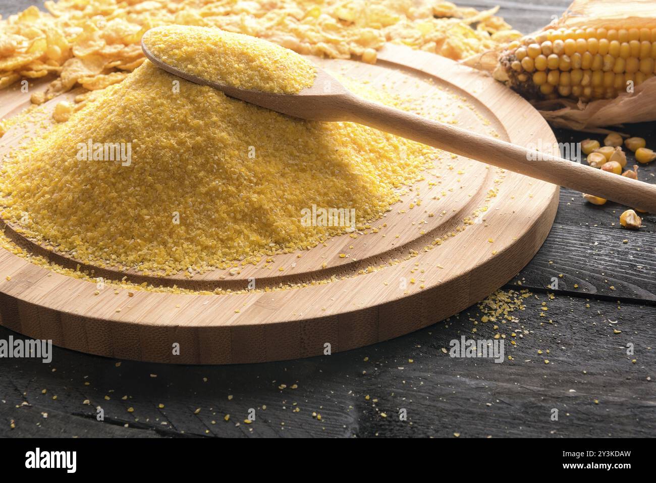 Cospargere di farina di mais su un piatto di legno con un cucchiaio di legno sopra di esso, circondato da pannocchie di mais, grani e fiocchi di cereali, su un backgroun di legno nero Foto Stock