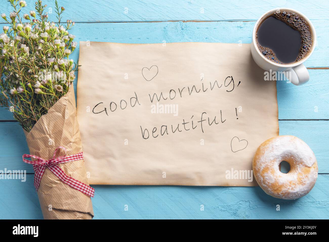 Carta d'epoca con il bel messaggio e i cuori del buon mattino, un bouquet di fiori primaverili, caffè caldo e ciambelle, su uno sfondo blu di legno Foto Stock