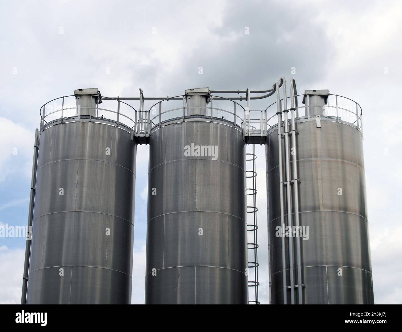 Un gruppo di tre serbatoi di stoccaggio di prodotti chimici in acciaio alti con tubi di collegamento e scale contro un cielo nuvoloso blu Foto Stock