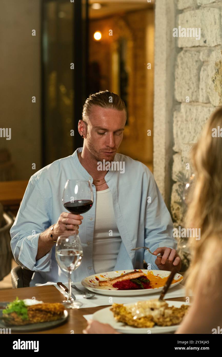 Un uomo assaggia attentamente il suo bicchiere di vino rosso mentre cena in un elegante ristorante all'aperto, assaporando i sapori Foto Stock