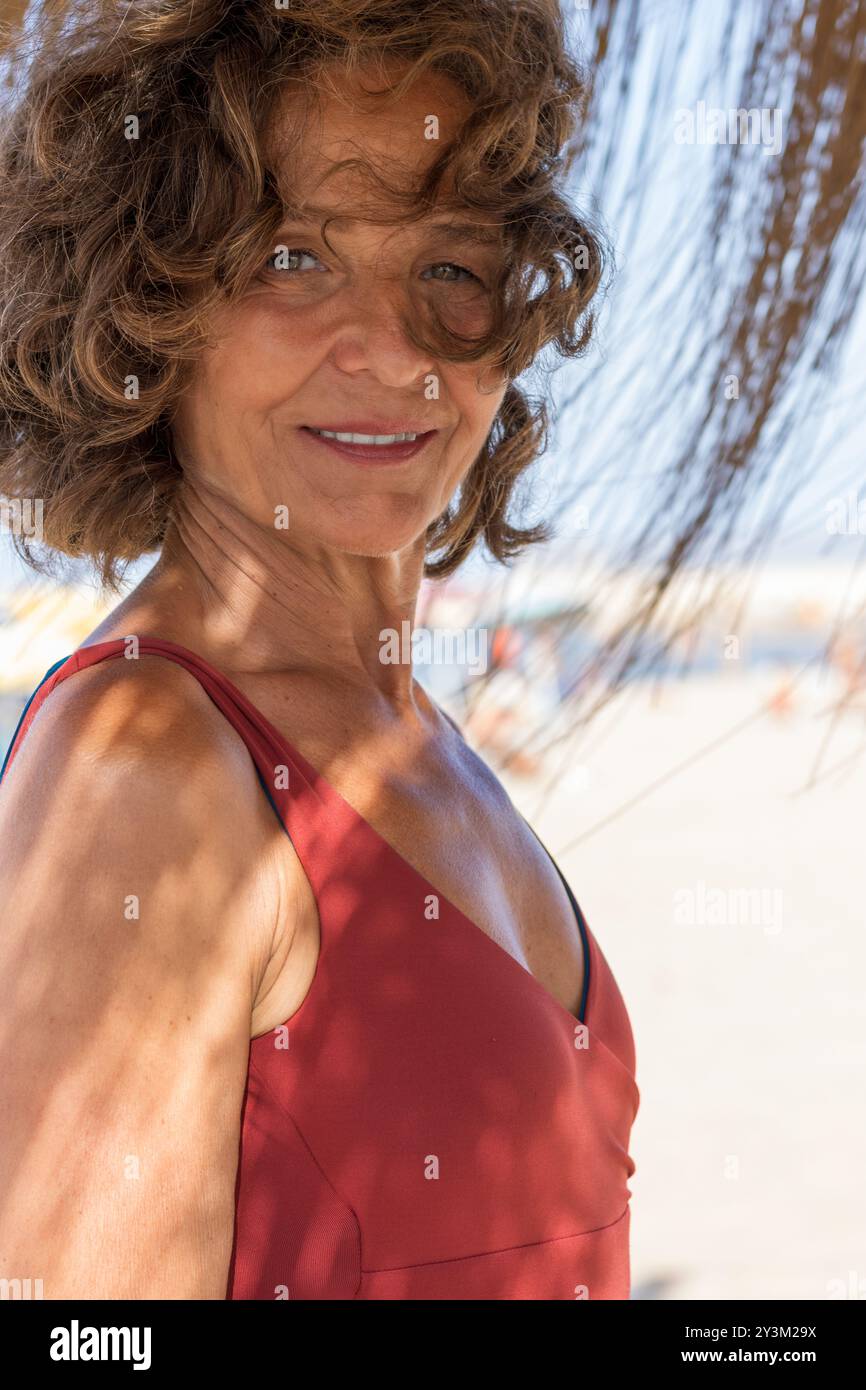 Una donna anziana è vista in piedi su una spiaggia di sabbia, con indosso un tetto senza maniche rosso. Gli elementi di fondo, tra cui sabbia e una foglia di palma, suggeriscono una t Foto Stock