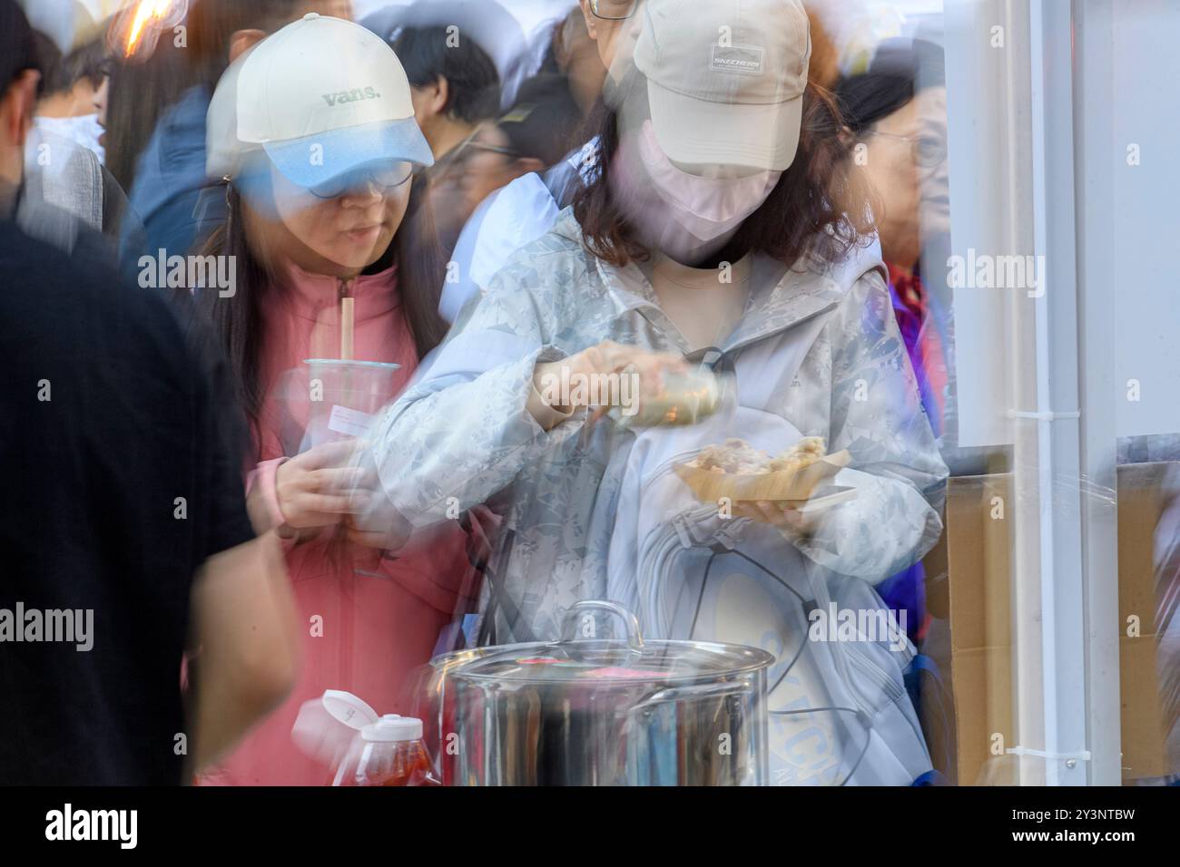 Due donne che indossano un cappellino stanno gustando il cibo di un venditore di Street food durante un evento di mercato molto affollato durante il giorno. Immagine sfocata. Calgary, Alberta Foto Stock