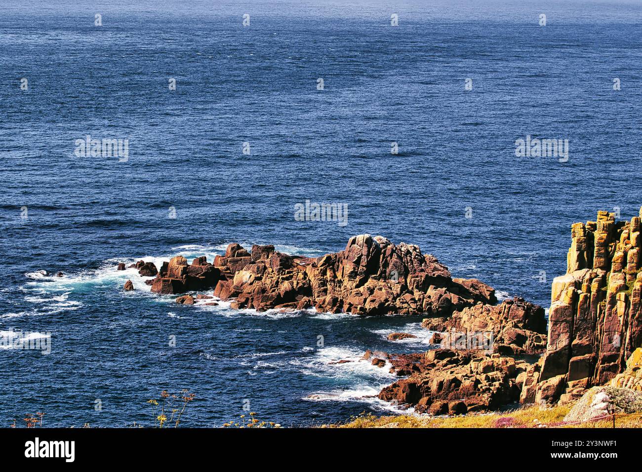 Una vista panoramica della costa rocciosa con onde che si infrangono contro le rocce, circondate da profonde acque blu dell'oceano. Le rocce mostrano vari colori e testo Foto Stock