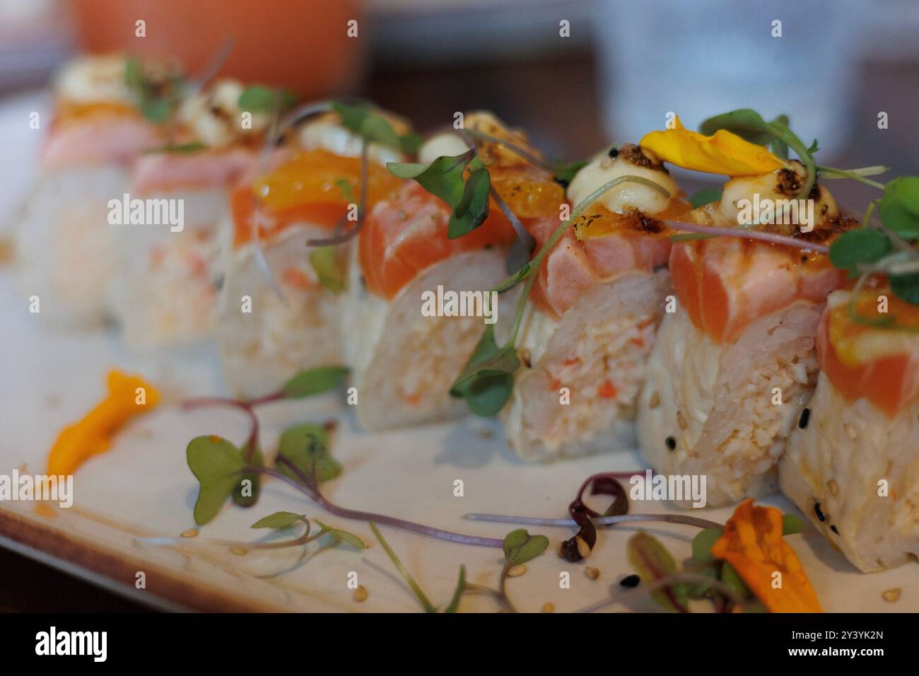 Primo piano di una specialità di sushi roll con salmone scottato, granchio piccante, tempura di gamberi e aioli, guarniti con microgreen, in mostra un'atmosfera vivace Foto Stock