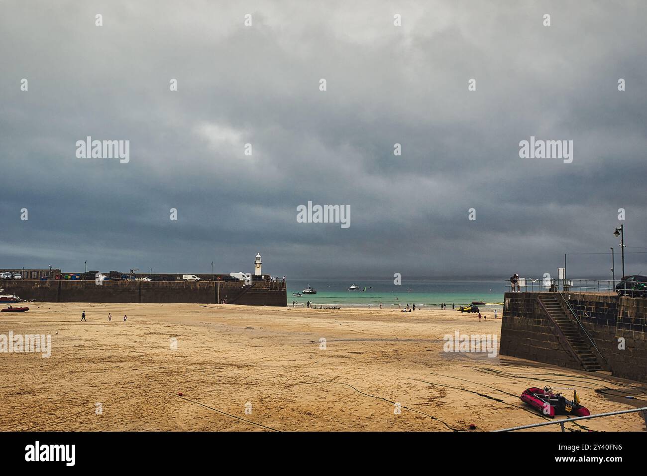 Una scena costiera caratterizzata da una spiaggia sabbiosa sotto un cielo nuvoloso. L'orizzonte mostra un mare calmo con barche in lontananza e un faro visibile. Persone a Foto Stock