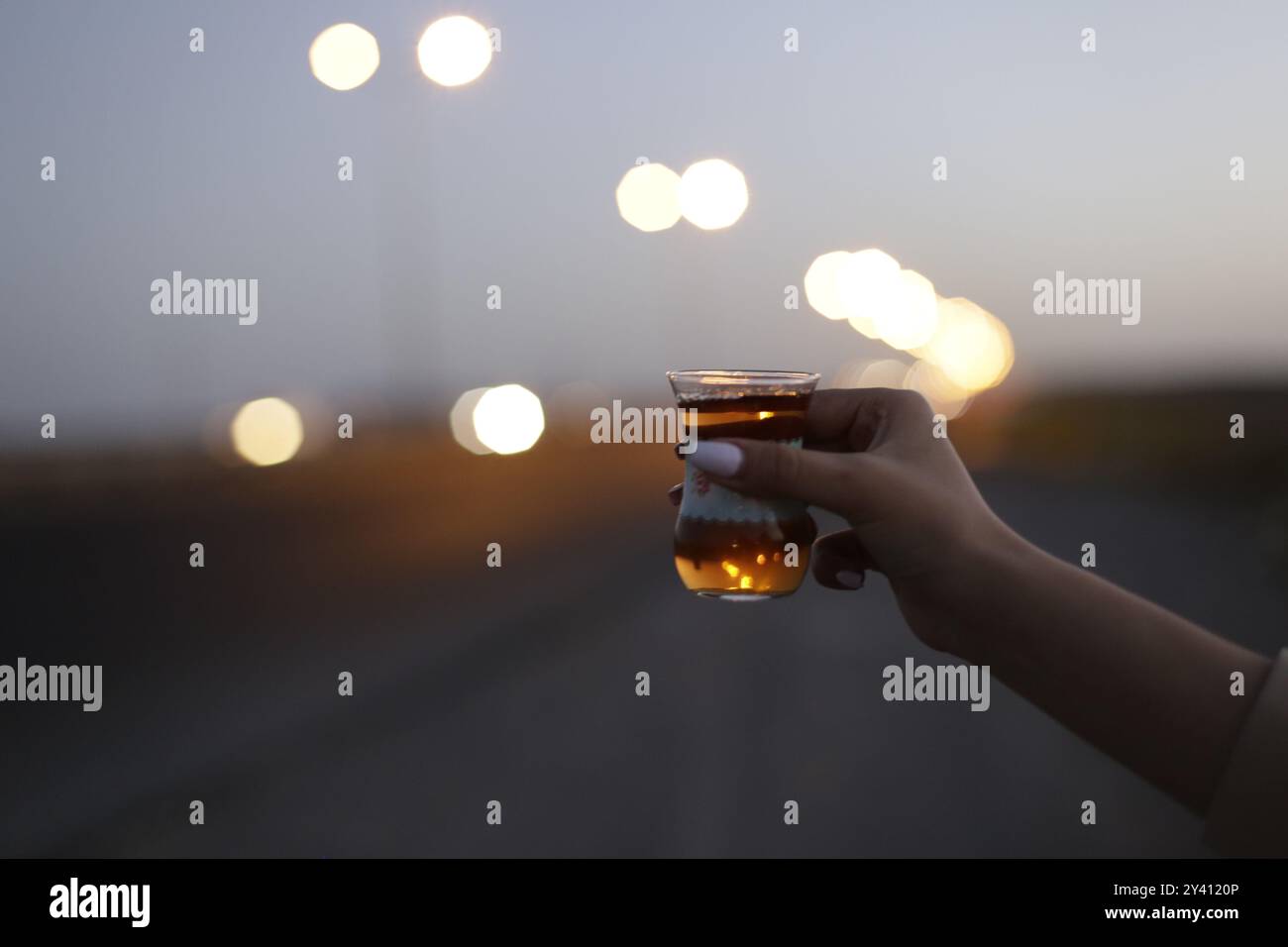 Una mano che regge un tradizionale bicchiere da tè con luci sfocate sullo sfondo, evocando un'atmosfera serena Foto Stock