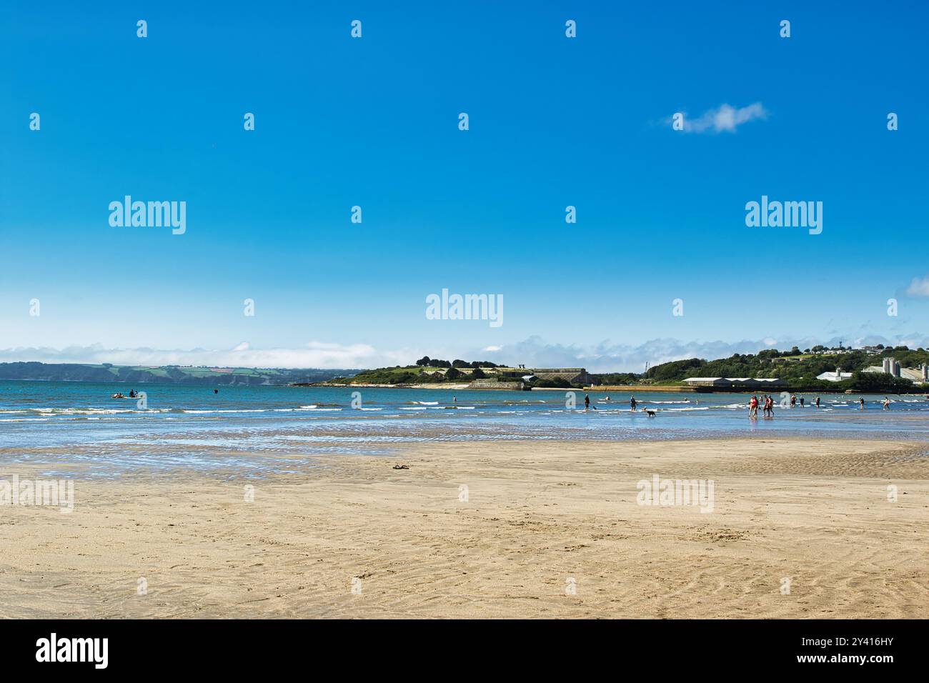 Una tranquilla spiaggia con sabbia dorata, onde dolci e un cielo azzurro. Le persone si godono varie attività in acqua, mentre verdi colline e. Foto Stock