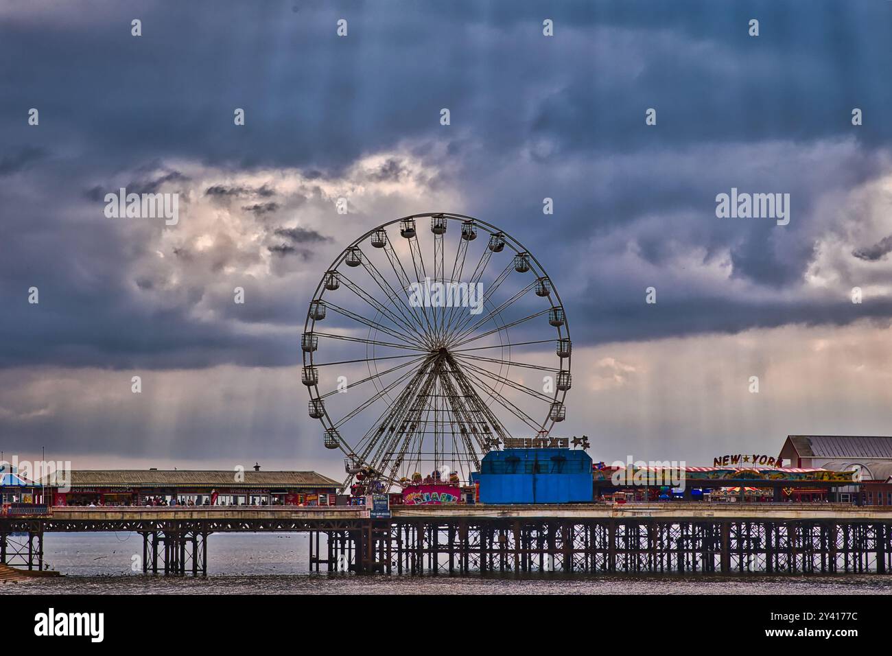 Una vista panoramica di una ruota panoramica su un molo sotto un cielo spettacolare con nuvole e raggi di luce solare. Il molo presenta attrazioni colorate e un vivace Foto Stock