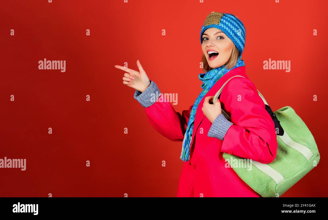 Donna sorridente con cappello blu, sciarpa e cappotto rosso con borsa a maglia verde che punta il dito sullo spazio copia. Moda primavera autunno inverno. Elegante ragazza dentro Foto Stock