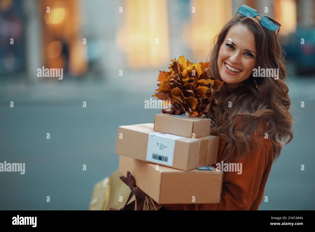 Ciao ottobre. donna elegante e felice in trench marrone con pacchi e foglie gialle autunnali in città. Foto Stock