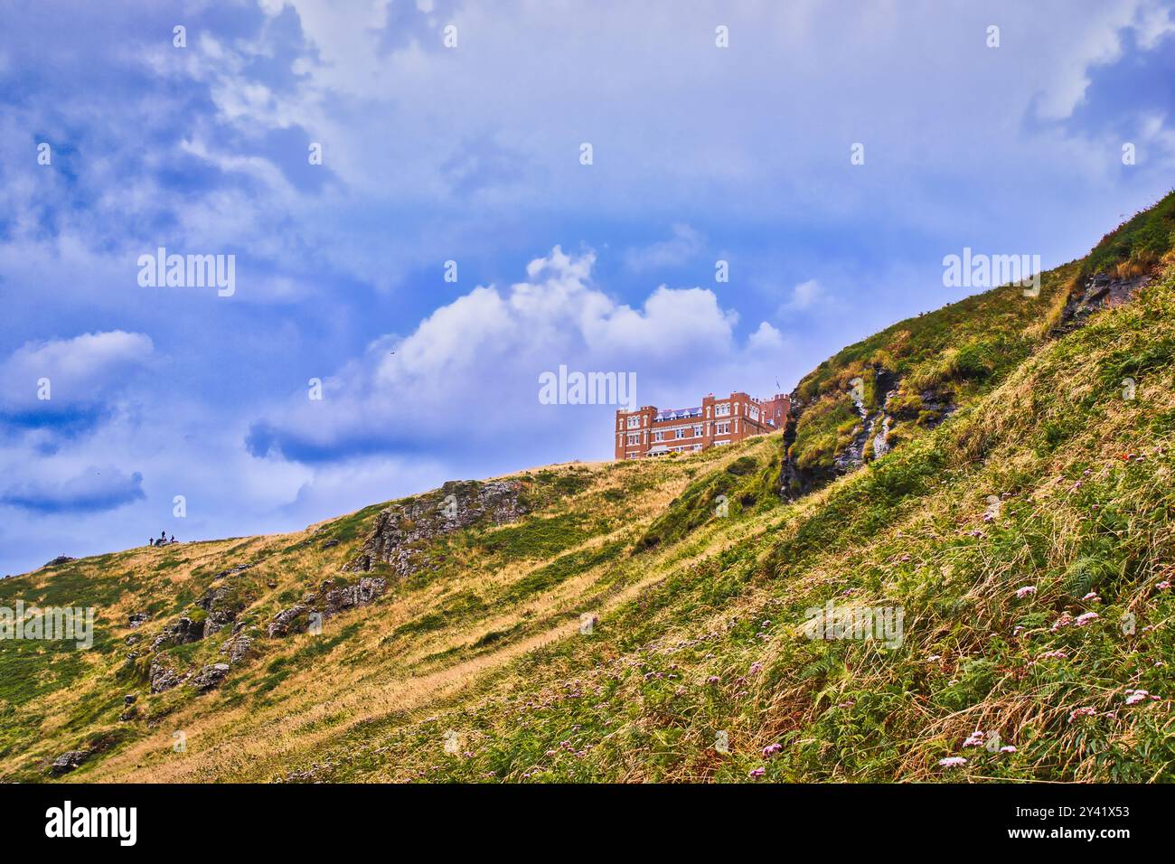 Una vista panoramica di una collina con lussureggiante erba verde e affioramenti rocciosi, caratterizzata da un edificio lontano arroccato sul pendio sotto un cielo parzialmente nuvoloso. Foto Stock