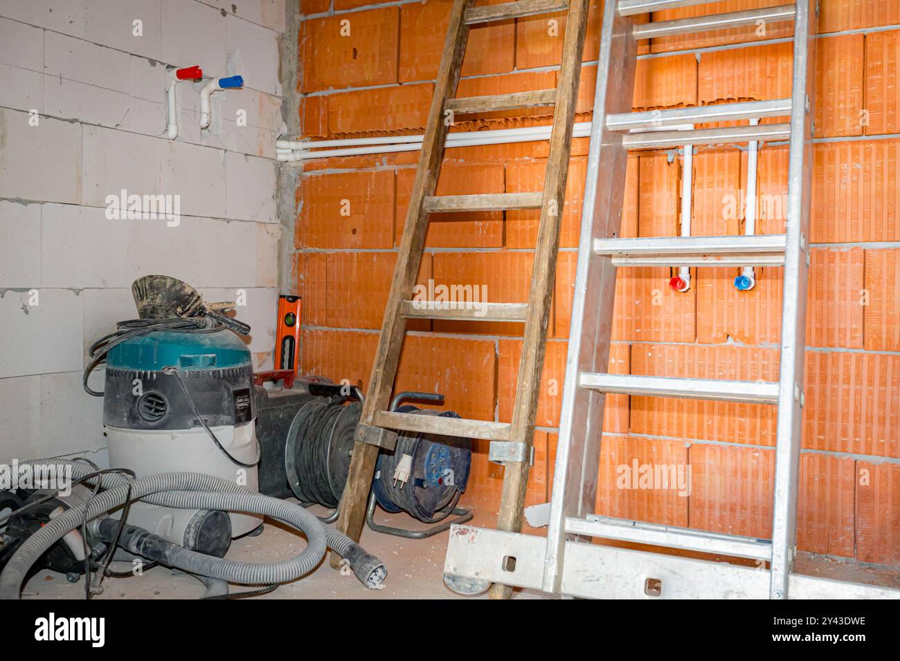 Scale in legno e metallo appoggiate contro pareti in blocchi rossi, collocate in un angolo del locale in costruzione, con aspirapolvere industriale e pochi aspirapolvere Foto Stock