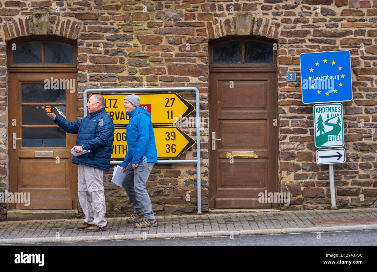 Segnale stradali gialli per Wiltz, Clervaux, Bastogne e St. Vith, con un indicatore del Lussemburgo e un cartello per sentieri, sono montati su un edificio in pietra, Lussemburgo. Foto Stock