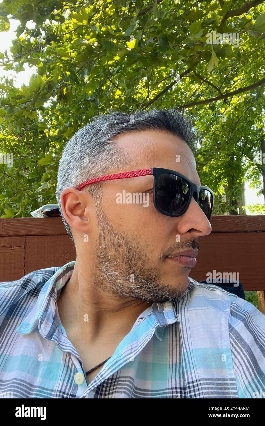 Un uomo che indossa occhiali da sole e una camicia a quadri. È seduto su una panchina. Gli occhiali da sole sono neri e rossi Foto Stock Un uomo che indossa occhiali da sole e una camicia a quadri. È seduto su una panchina. Gli occhiali da sole sono neri e rossi Foto Stock