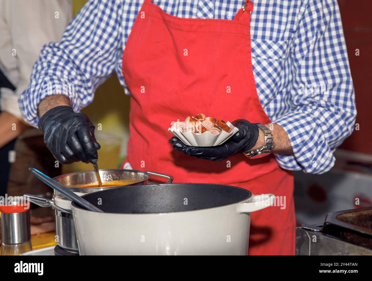 Chef vestito con grembiule rosso, con fette di salsiccia, ingredienti placcati, preparazione di un piatto delizioso in una cucina professionale, arte culinaria c Foto Stock