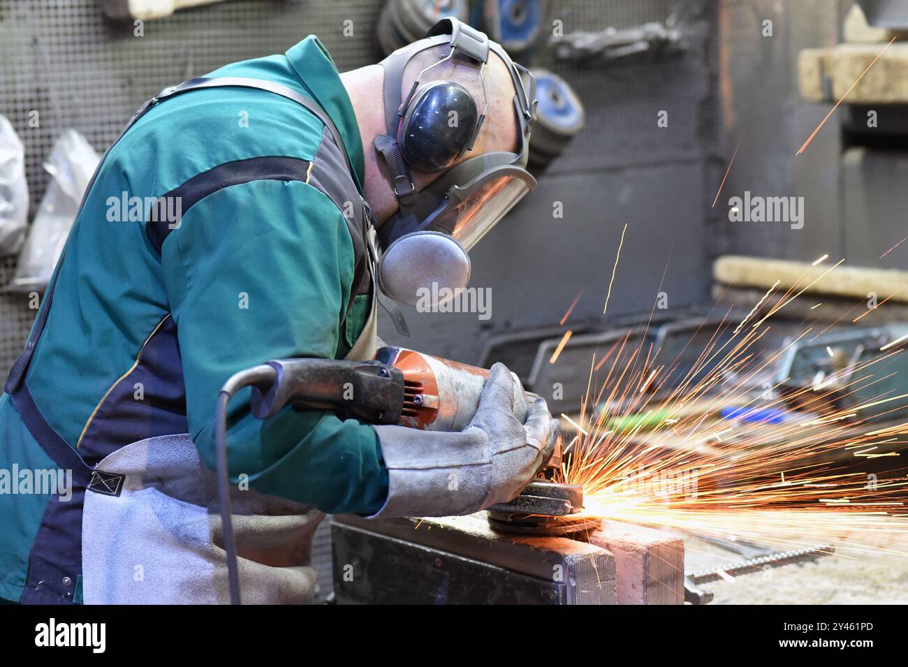 Il lavoratore con una rettificatrice lavora un pezzo in acciaio in un'azienda industriale - sicurezza sul lavoro con protezione per orecchie e occhi - foto ravvicinata Foto Stock