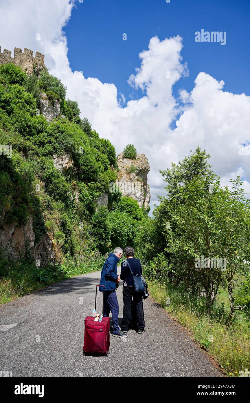 Due viaggiatori con una valigia si fermano su una strada panoramica circondata da vegetazione lussureggiante e scogliere ripide, con il castello di Óbidos visibile sulla cima della collina Foto Stock