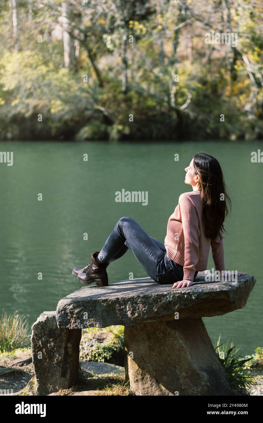 Donna che si gode il sole invernale, circondata dal verde e dalle acque tranquille, seduta su una panchina di pietra sul fiume Eume in Galizia, Spagna Foto Stock