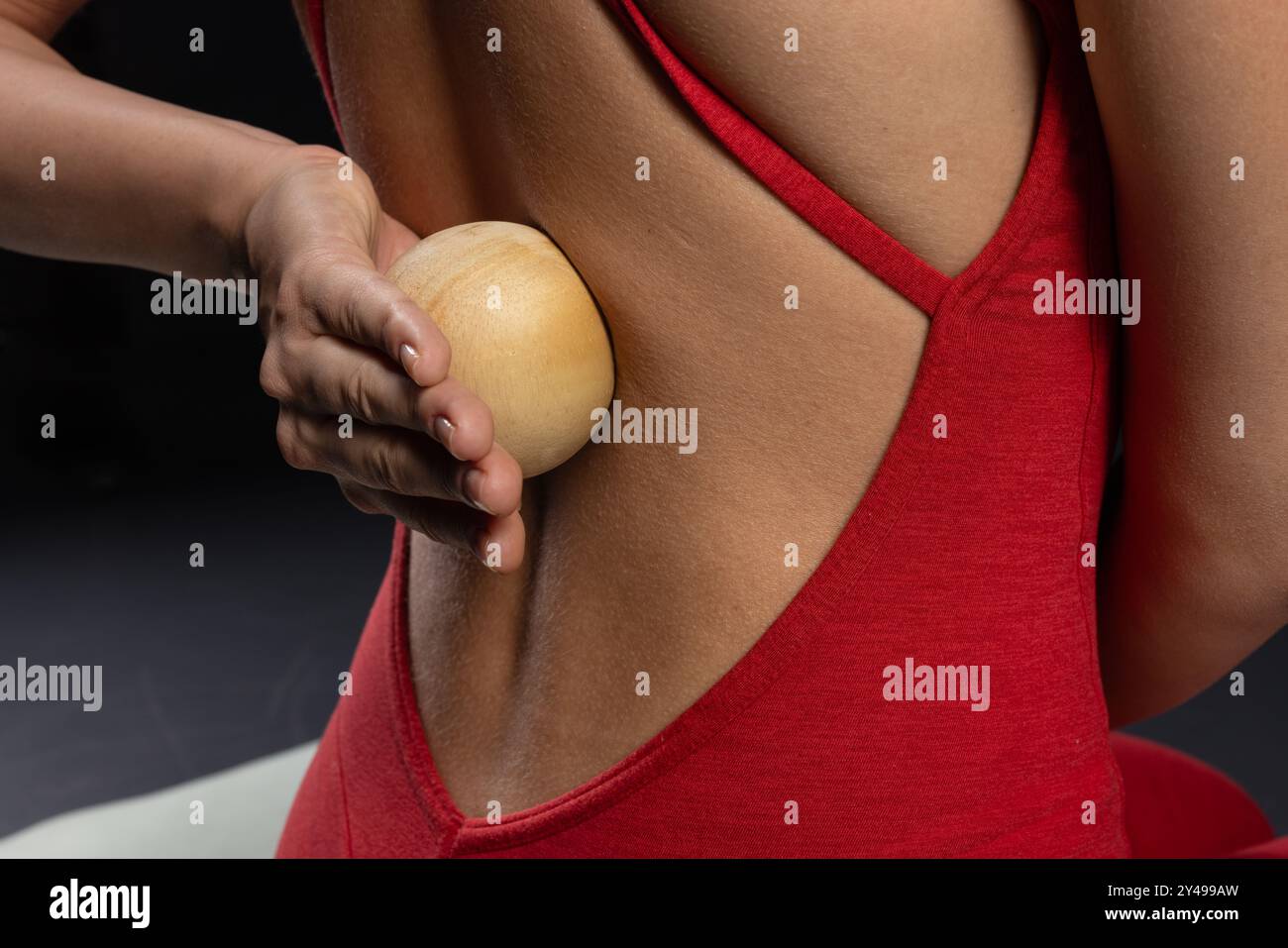 ragazza che fa massaggio muscolare alla schiena con palla da yoga in legno in maglietta rossa su sfondo scuro, cura di sé e tecniche di rilassamento. strumento di auto-massaggio Foto Stock