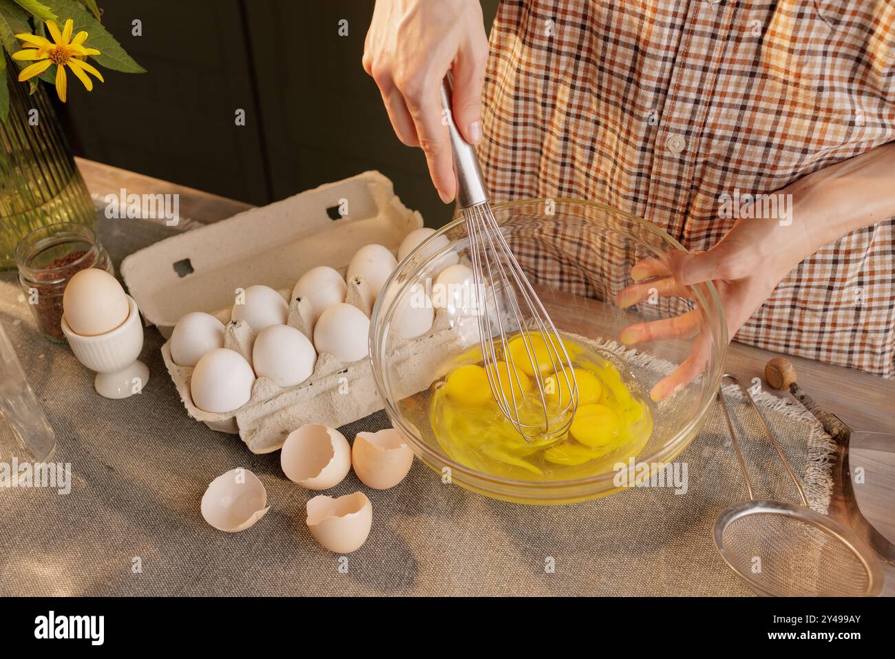 Primo piano di mani femminili che battono le uova con una frusta in un recipiente trasparente sullo sfondo di un tavolo da cucina. Uova in una scatola e uno strumento per Foto Stock