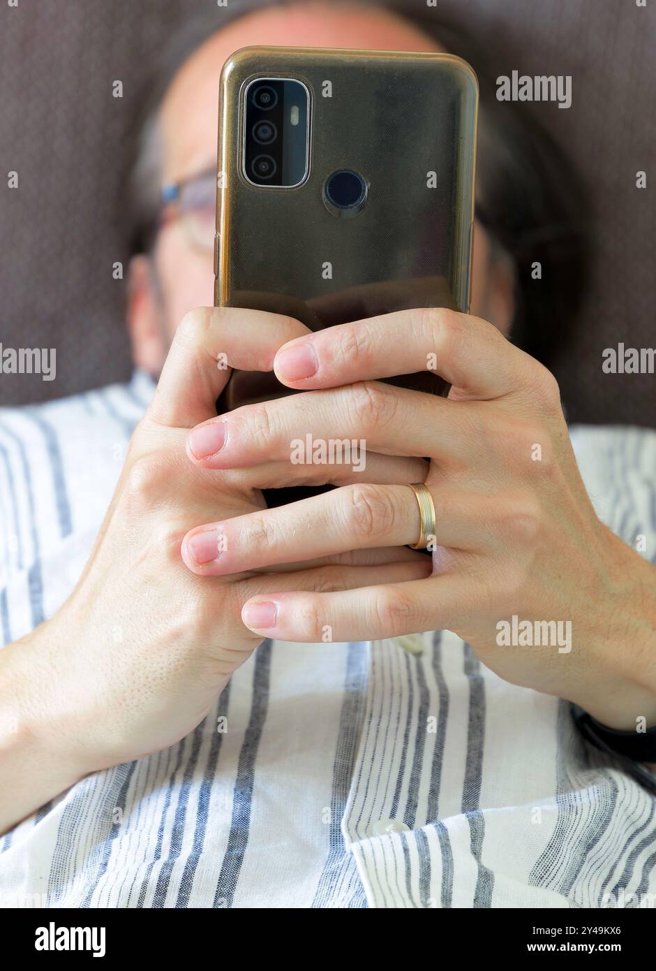 Un uomo sta guardando il suo telefono mentre si stende su un divano. Indossa un anello nuziale e il telefono gli copre la faccia Foto Stock