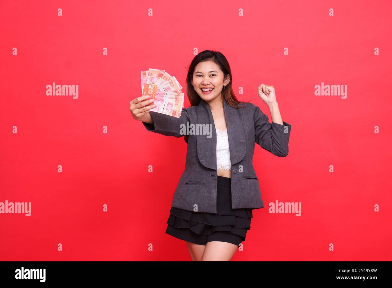 Espressione di una donna asiatica felice in carriera che tiene una carta di credito e denaro davanti a un cartello che stringe il pugno a lato con giacca e gonna Foto Stock