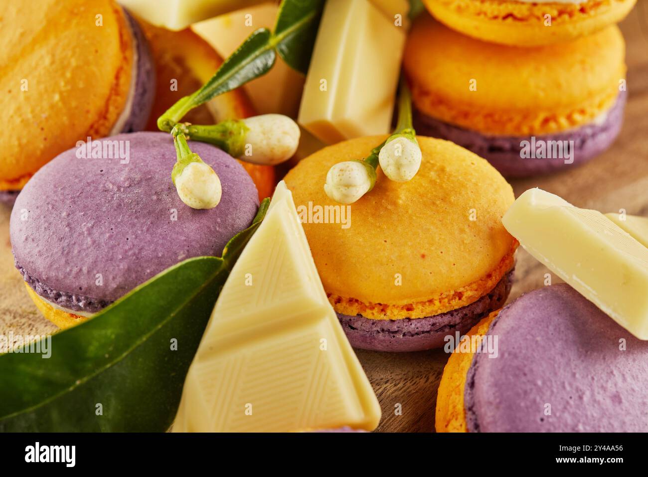 Vivaci macaron gialli e viola visualizzati con fette arancioni, foglie verdi e pezzi di cioccolato bianco su una tavola rustica di legno. Foto Stock