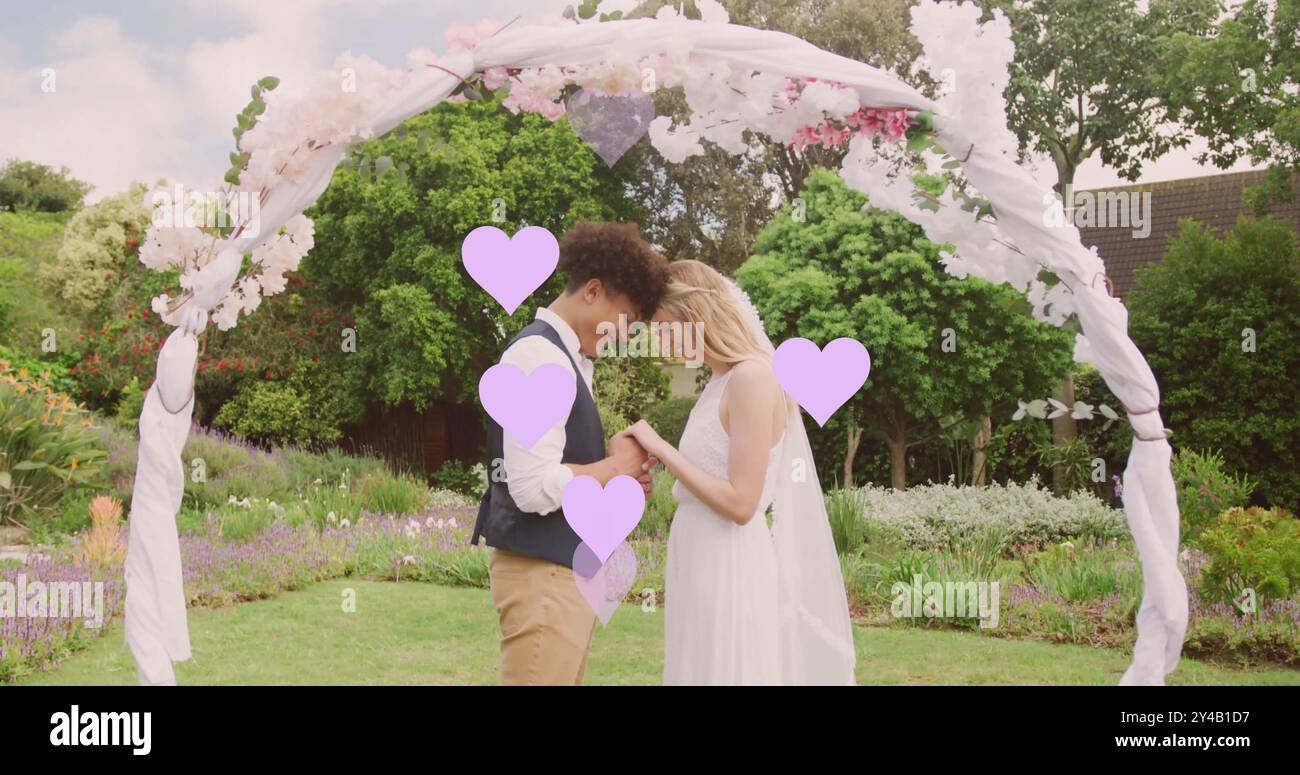 Immagine di cuori lilla e di una coppia felice e variegata che tiene per mano sotto l'arco nuziale in giardino Foto Stock