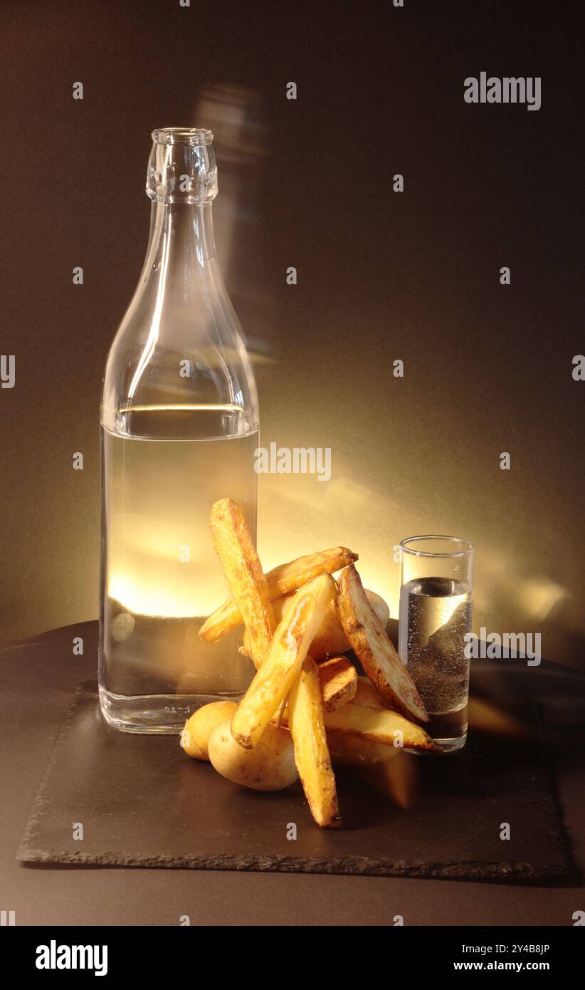 Patate rustiche accanto a un bicchiere e bicchierino pieno di liquido trasparente. La luce gialla crea un triangolo intrigante sullo sfondo scuro. Foto Stock