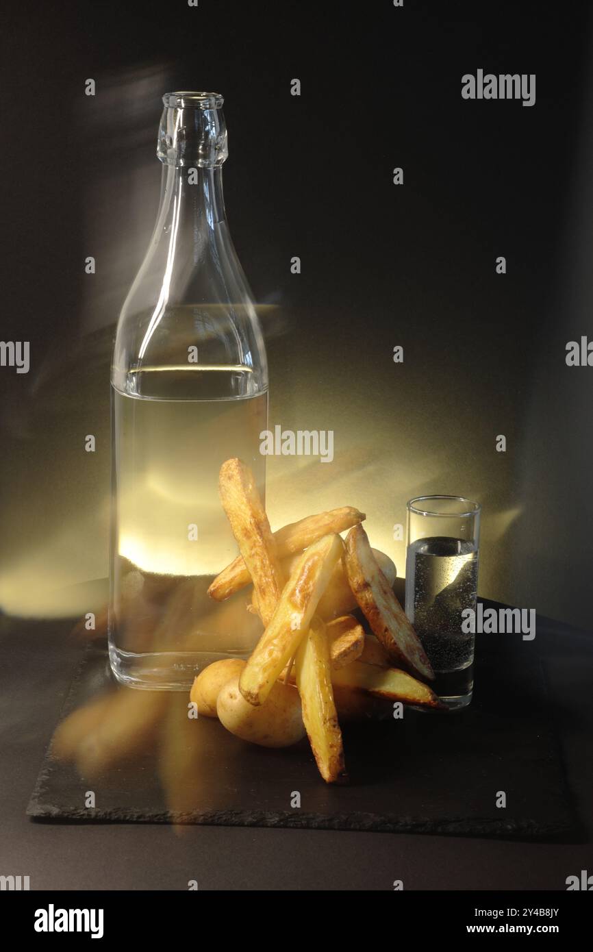 Un bicchiere e un bicchierino di liquido limpido accanto alle patate rustiche su sfondo scuro. Il triangolo di luce aggiunge calore e contrasta l'ambiente buio Foto Stock