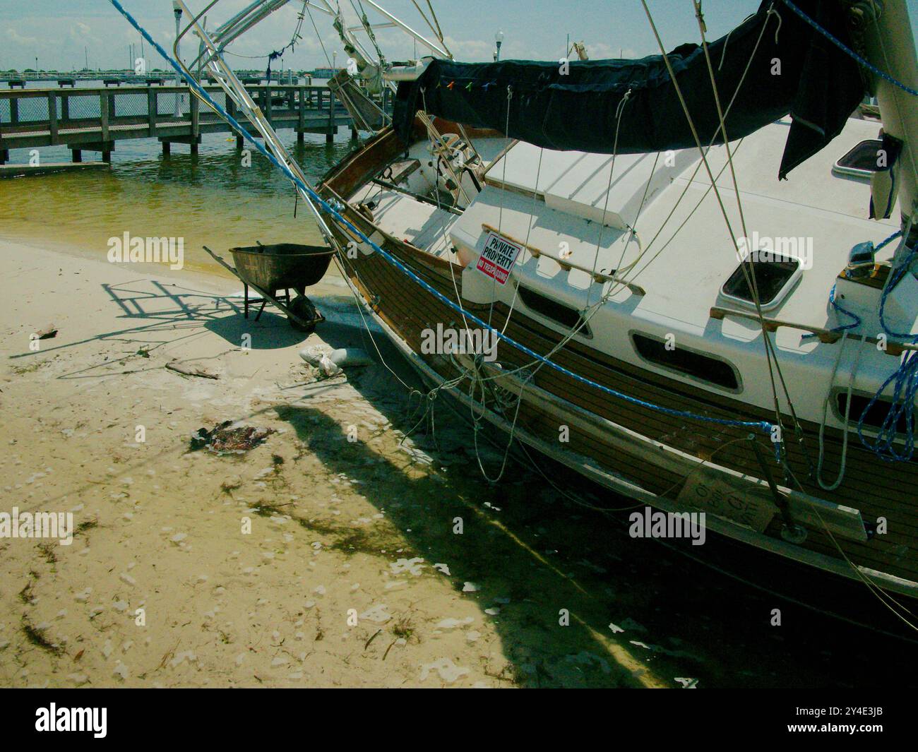 Barca a vela bianca e azzurra danneggiata appoggiata a destra sulla spiaggia tra alberi dopo una tempesta a Gulfport, Florida. Guarda dalla spiaggia alla baia Foto Stock