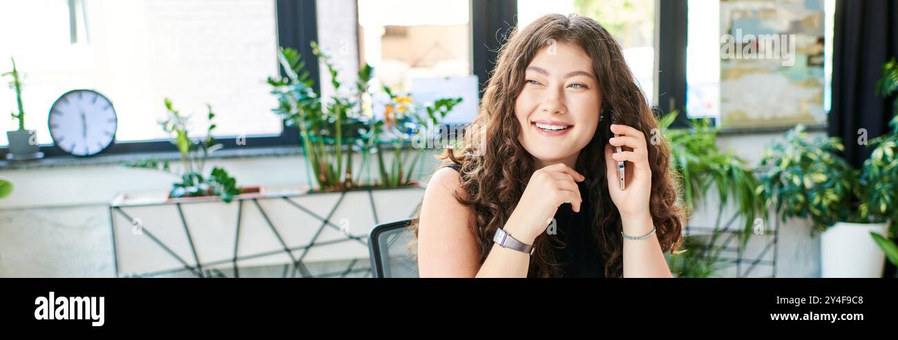 Una giovane donna di grandi dimensioni sorride brillantemente mentre parla al telefono, circondata da vivaci piante d'ufficio. Foto Stock