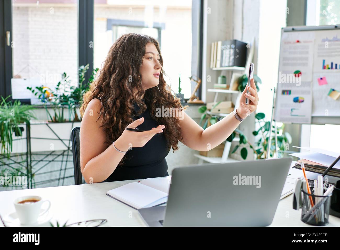 Una giovane donna d'affari con i capelli ricci conduce una videochiamata mentre è circondata da piante e un'invitante area di lavoro. Foto Stock