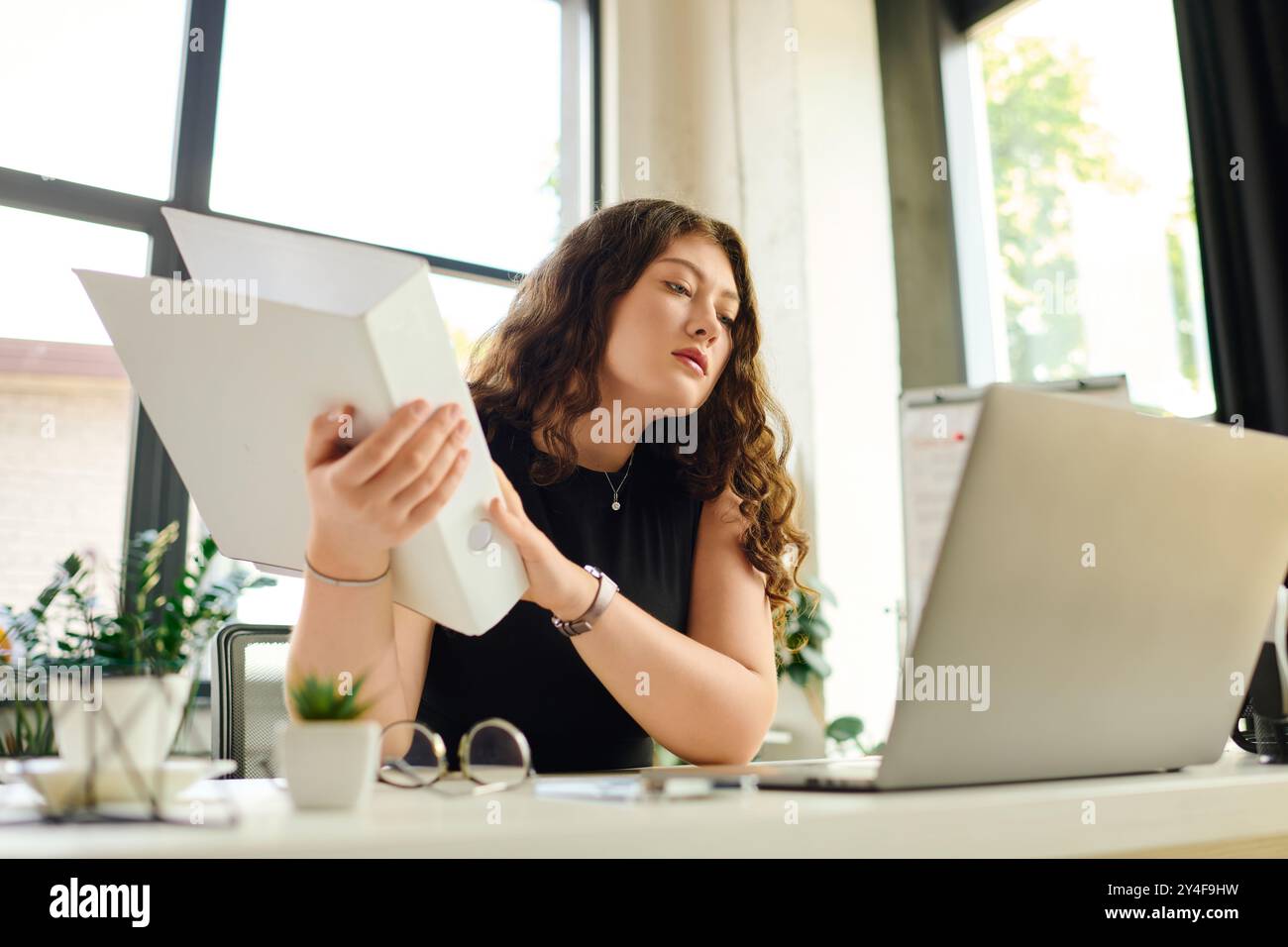 Una donna giovane, bella e di grandi dimensioni con lunghi capelli ricci si concentra intensamente sui documenti mentre lavora alla scrivania in ufficio. Foto Stock