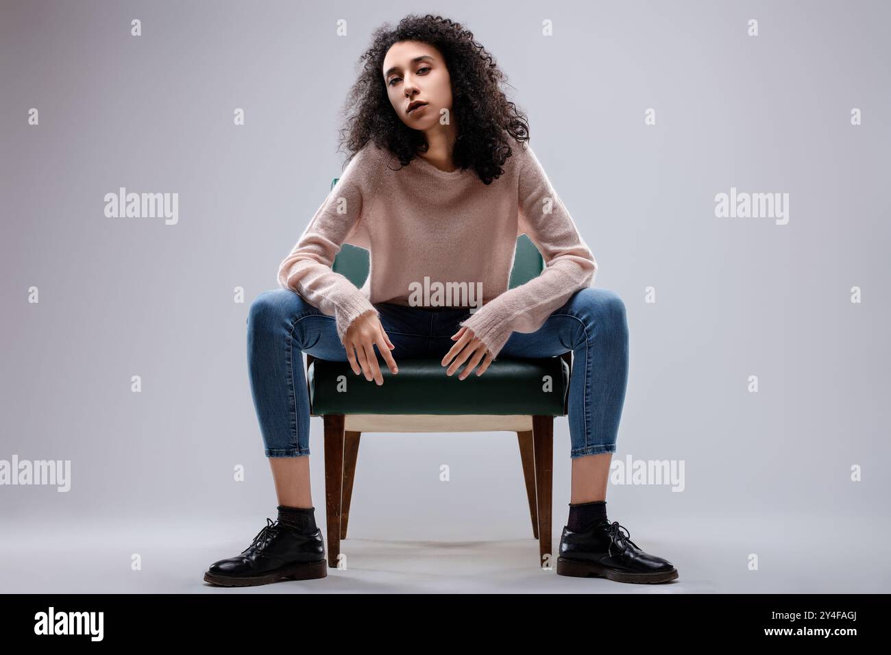 Modella di moda con capelli ricci e maglione rosa che si posa seduta su una sedia verde, allarga le gambe e poggia le mani sulle cosce Foto Stock