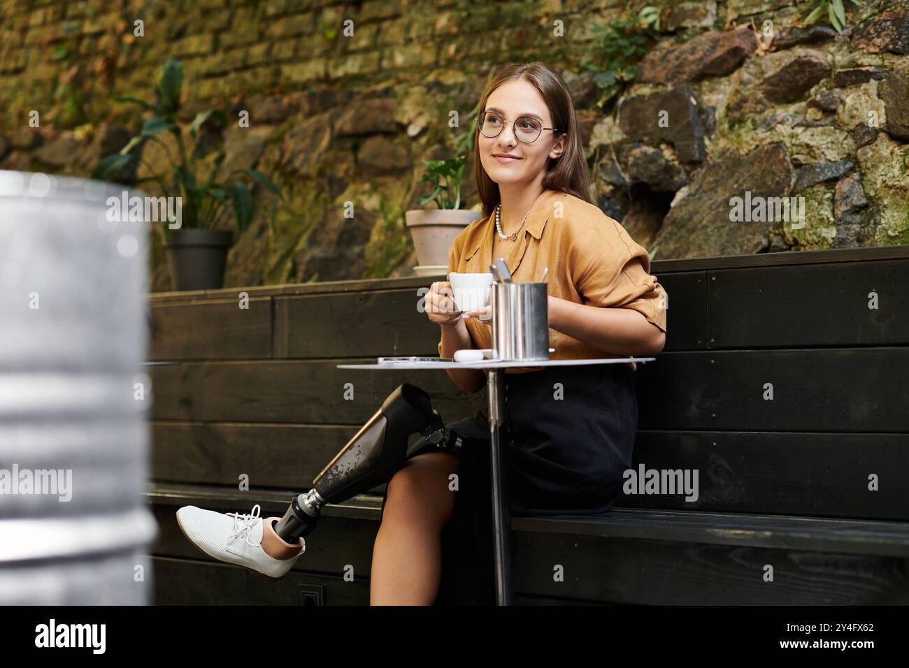 Una giovane donna siede comodamente in un caffè, assaporando il caffè mentre abbraccia il suo stile di vita unico con una gamba protesica. Foto Stock