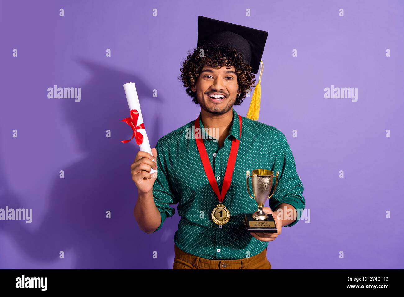 Foto di un giovane studente indiano laureato in nappina che tiene il suo diploma medaglia d'oro e calice isolato su sfondo di colore viola Foto Stock