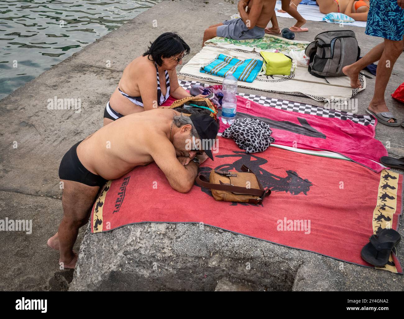 Un uomo italiano in costume da bagno dorme in piedi e riposa su un asciugamano Ferrari sulla banchina di Castro Marina, Puglia, Italia. Foto Stock