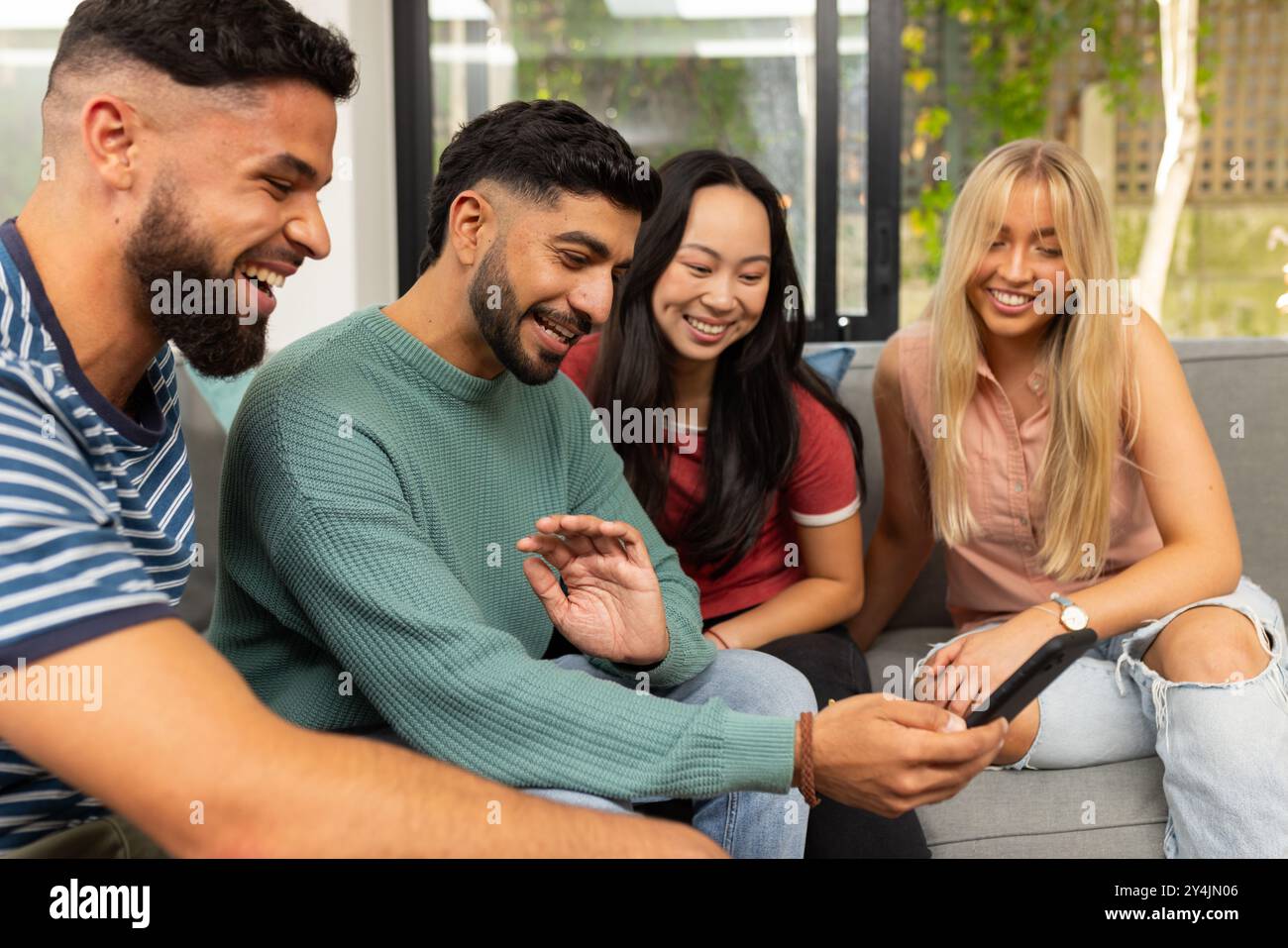 amici diversi seduti sul divano, sorridono e guardano insieme lo smartphone Foto Stock
