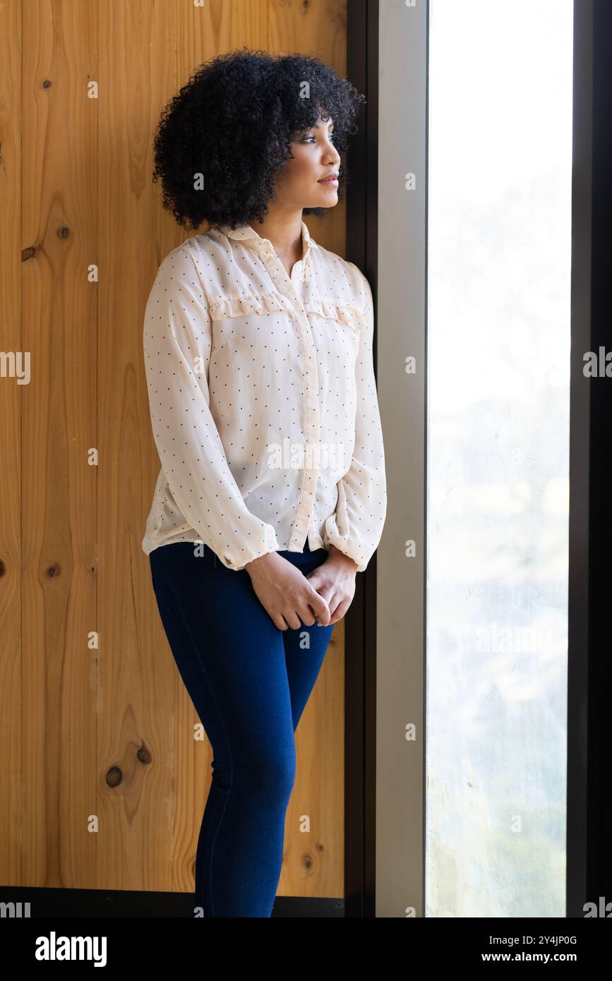 In piedi accanto alla finestra, donna in un abbigliamento informale che guarda all'esterno con attenzione Foto Stock