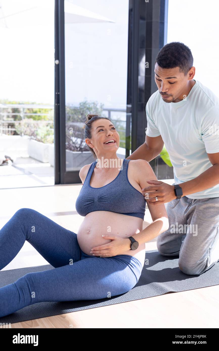 A casa, coppia multirazziale, donna incinta che si esercita su un tappetino yoga con assistenza partner Foto Stock