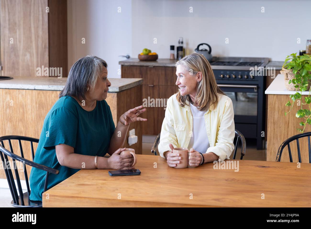 A casa, amiche anziane multirazziali sedute a tavola, bevendo un caffè e chiacchierando in cucina Foto Stock